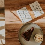 A cozy café setup with cake, ice cream, and a book on a sunlit wooden table.